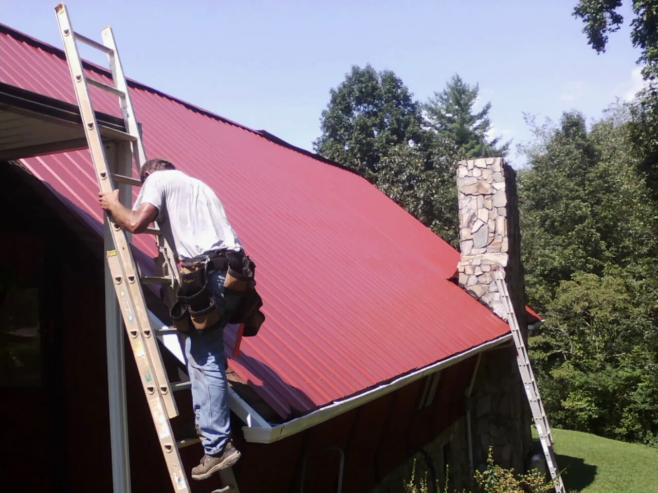 Roofer on a ladder beside a red metal roof and stone chimney — Soffit & Fascia Repair by Roofing Brothers in Coalmont